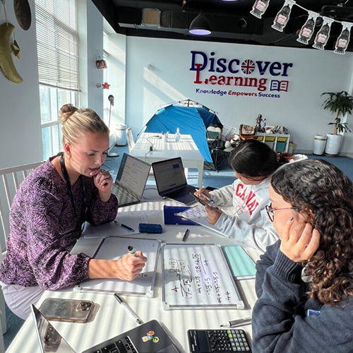 Three students collaborate at a bright classroom table, one writes math on a whiteboard while two work on laptops; a blue tent and the 'Discover Learning' slogan are in the background.