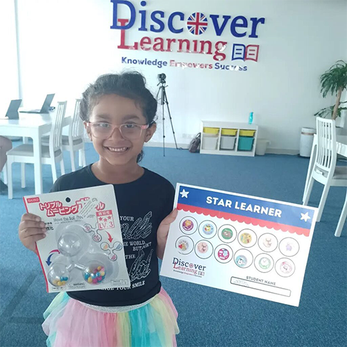 Smiling girl in a Discover Learning classroom holds a bubble toy package and a Star Learner certificate.