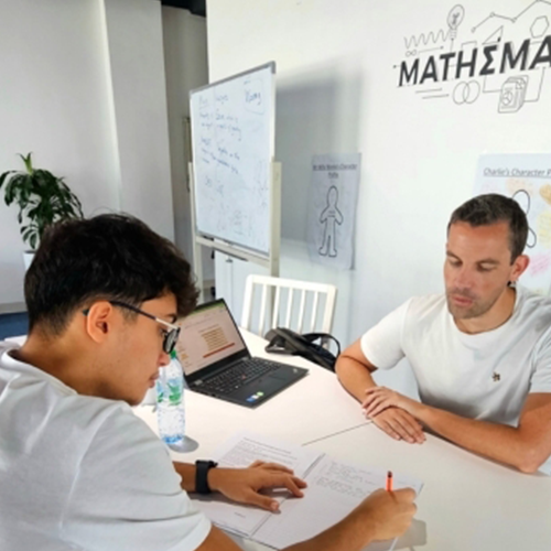 Two men at a table in a math tutoring room; one writes on paper as the other watches, with a laptop nearby.