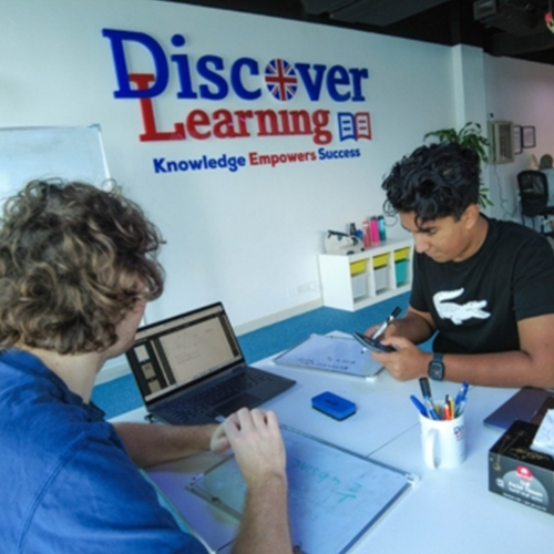 Two students study at a Discover Learning center; one uses a laptop while the other writes on a notebook at a white desk, with the center's logo and slogan on the wall.