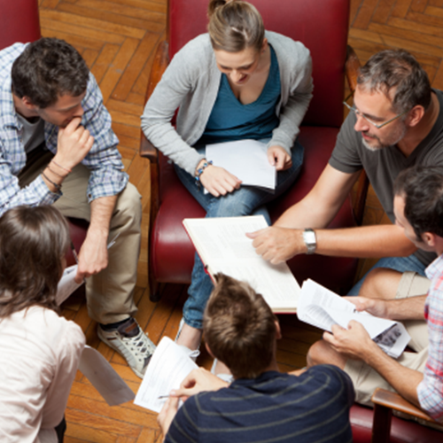 Six adults seated in a circle, reading documents and discussing together in a casual meeting space.
